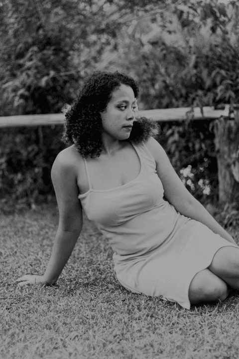 leticia sentada na grama, com uma mão sobre o corpo e outra na grama, olhando para o lado, com vegetação e o lago ao fundo da imagem'