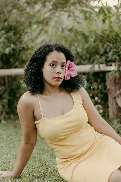 leticia sentada na grama, com uma mão sobre o corpo e outra na grama, olhando para mim, bem bonita'