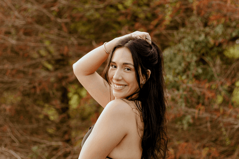 lu segurando cabelo em um penteado ao alto da cabeça, olhando para a câmera, sorrindo, com as árvores avermelhadas de fundo'
