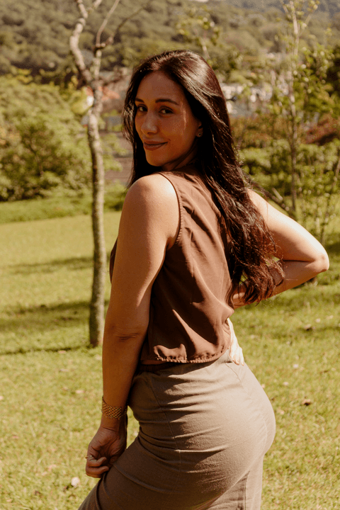 luiza com uma mão na cintura, mais de costas e lateral para mim, com um sorriso leve, natureza ao fundo'