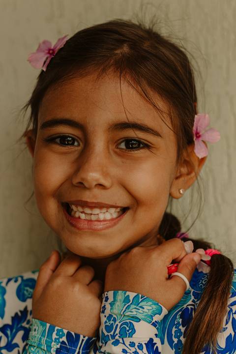 maria alice com sorriso largo, flores em seu cabelo, maiô azul e branco, com as mãos próximas ao seu rosto'