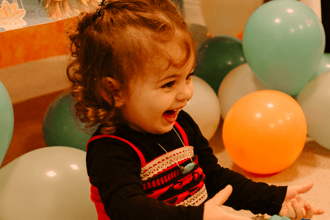 maria eduarda sorrindo aos montes em frente a mesa de decoração do seu aniversário, com balóes ao fundo'