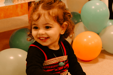 maria sorrindo sentada em frente a mesa de decoração e bolo da festa'
