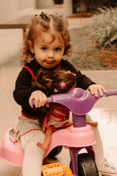 maria eduarda na sua motoca, com uma boneca da moana no colo, mostrando a língua'