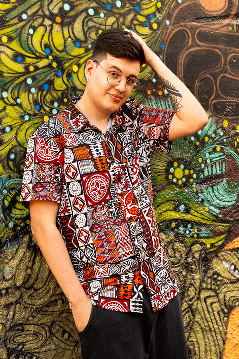 nathan em frente a parede colorida, com sua blusa também colorida, passando a mão no cabelo enquanto sorri'