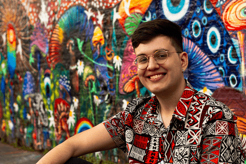 nathan sentado na calçada em frnete a parede grafitada, sorrindo'