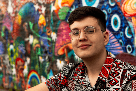 nathan sentado na calçada em frnete a parede grafitada, leve sorriso em seu rosto, mais próximo'