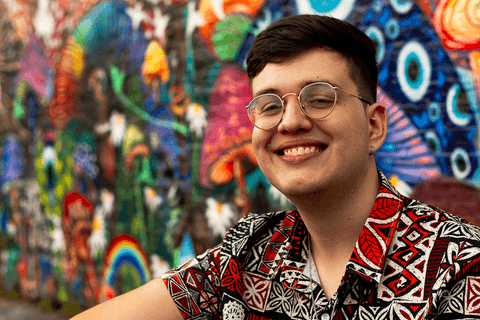 nathan sentado na calçada em frnete a parede grafitada, sorrisão bem aberto em seu rosto, mais próximo'