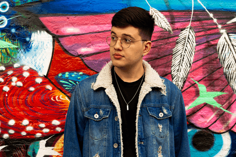 nathan olhando para o horizonte, com jaqueta jeans, em frente a parede grafitada'