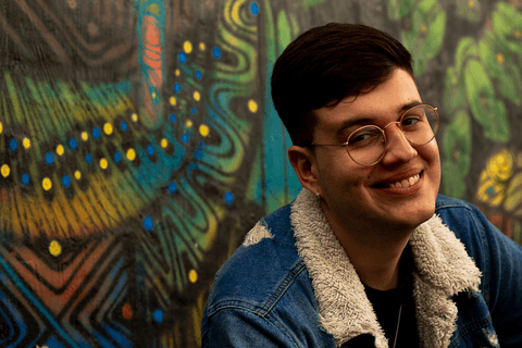 nathan sentado em frente a parede grafitada, com sorriso no rosto'