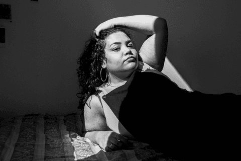 suelen com uma das mãos sobre seu cabelo, olhando para mim, deitada em sua cama, luz do sol pegando partes do seu corpo'