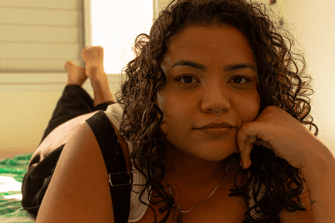 suelen deitada de barriga para baixo, com as pernas cruzadas e os pés levantados, com a mão apoiada no rosto, olhando para mim com um leve sorriso em seu rosto'