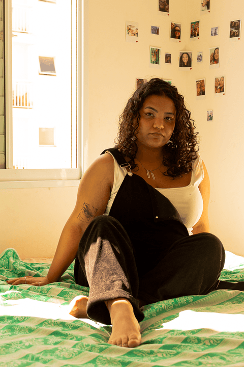 suelen sentada em sua cama, com as pernas cruzadas e uma das mãos apoiando na cama'