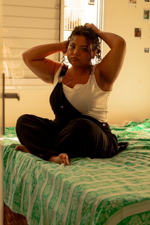 suelen amarrado seu cabelo, sentada de pernas cruzadas na sua cama, olhando para mim, janela de fundo com bastante luz do sol entrando no quarto'