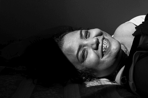 suelen deitada em sua cama, olhando para mim, luz do sol batendo em parte do seu rosto, com sorriso bem aberto'