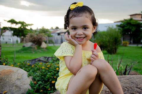 Ensaio pré aniversário de 03 anos - Nicole - Criança de 03 anos em sessão fotográfica - Criança sentada sobre uma pedra com a mão sob o queixo e sorrindo'
