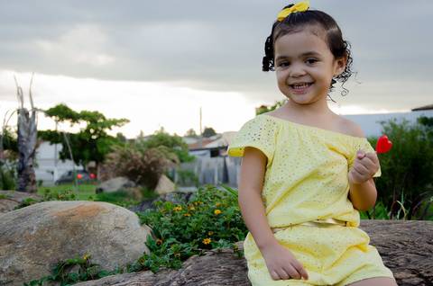 Ensaio pré aniversário de 03 anos - Nicole - Criança de 03 anos em sessão fotográfica - Criança sentada sobre uma pedra com um pirulito vermelho na mão'