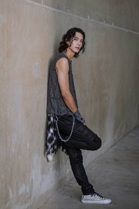 Plano inteiro de homem com visual rocker encostado em parede de concreto longa, usando calça preta com correntes e camisa xadrez na cintura.'