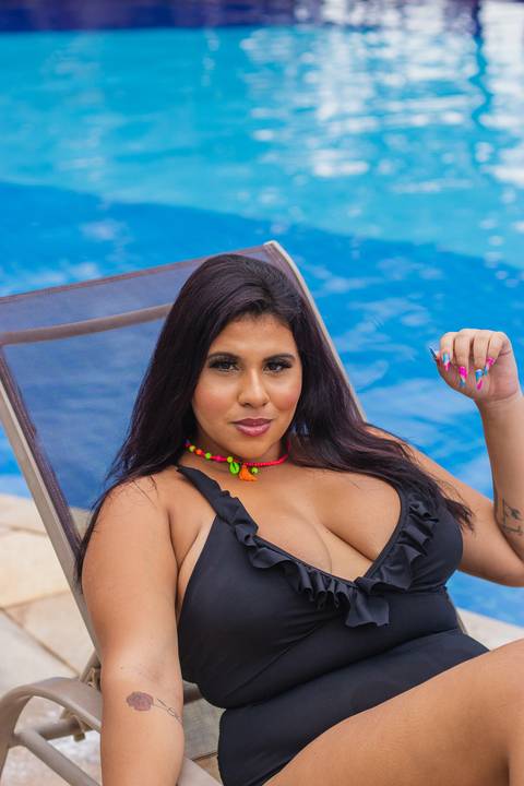 modelo plus size jovem de biquini preto e cabelo liso posa para foto em ensaio fotográfico feminino deitada em cadeira ao lado da piscina'