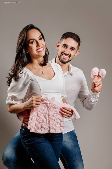 Fotografia de gravida segurando paletozinho de tricot com marido segurando sapatinhos em estúdio fotográfico'