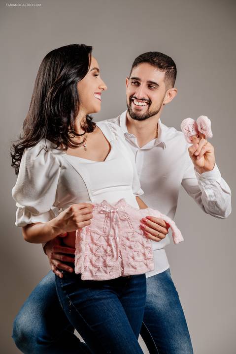 Fotografia de GeFotografia de gravida segurando paletozinho de tricot com marido segurando sapatinhos em estúdio fotográficostante com marido em estúdio fotográfico'