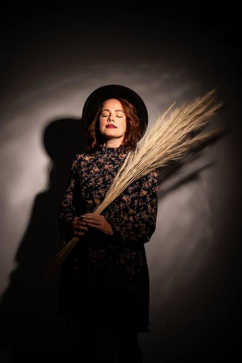Mulher segurando capim desidratado usando chapéu pretos e olhos fechados para a luz.'