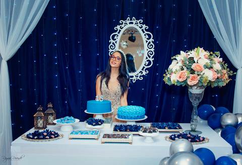 a mesa do bolo toda decorada na cor azul do jeito que a debutante gosta com bolos azuis e docinhos azuis com arranjo de flores alto e cortinas laterais brancas e um espelho no fundo e lamparinas como decoração'