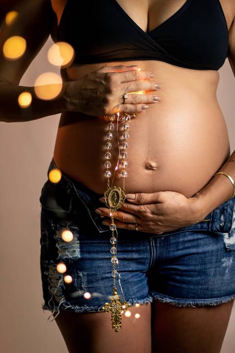 gestante segurando colar de cruz e luzes em sua barriga '
