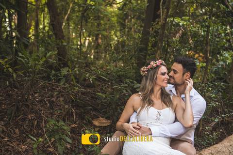 Casal sentado em um tronco de arvore a noica vestindo vestido casual branco co tiara de flores do campo, noivo com camisa branca, os dois quase se beijando, ela com  a mão no pescoço do noivo'