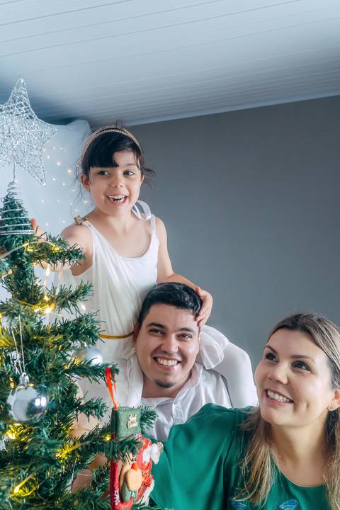 Ensaio de Natal em Família em Piraquara e Curitiba Paraná. Casal com sua filha enfeitando uma árvore de natal com luzes e enfeites.'