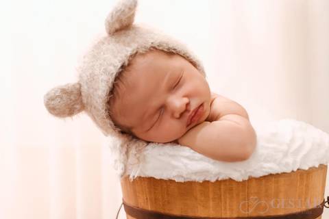 Bebê recém nascido com touca de ursinho na cor bege, dormindo em um baldinho de madeira.'