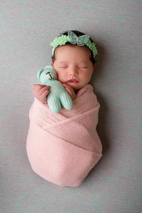 Foto da bebê Helena durante o ensaio newborn feito no estúdio Carol Zimmermann em São José, SC. Ela está enrolada num wrap cor de rosa e segurando um urinho de tricô verde claro.'