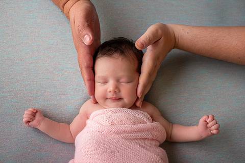 Bebê Helena no seu ensaio newborn, com o aconchego das mãos de sua mãe e de seu pai. Um sorriso no rosto mostra a felicidade da bebê ao sentir a presença dos pais ao seu lado. Foto pela fotógrafa Carol Zimmermann no estúdio em São José, SC.'