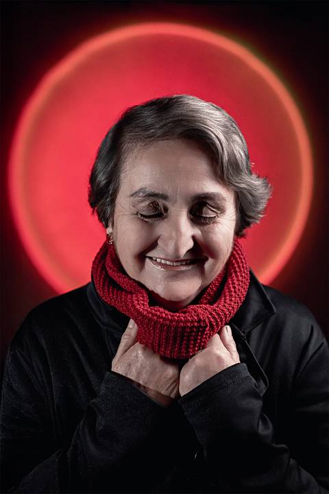 Imagem de mulher na faixa dos 50 anos de frente para câmera, olhos fechados e sorrind usando uma jaqueta preta.'