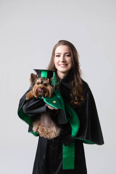Retrato oficial de formatura em Medicina. Maria Eduarda vestindo beca tradicional, capelo e segurando o diploma da faculdade.'