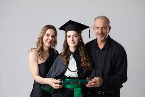 Retrato oficial de formatura em Medicina. Maria Eduarda vestindo beca tradicional, capelo e segurando o diploma da faculdade.'