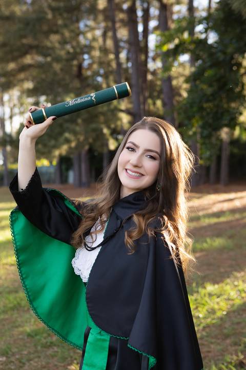 Retrato oficial de formatura em Medicina. Maria Eduarda vestindo beca tradicional, capelo e segurando o diploma da faculdade.'