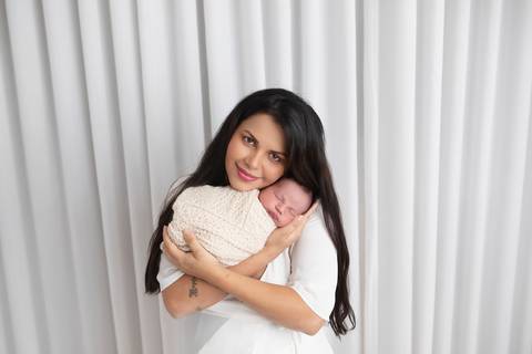 Fotografia afetiva de mãe e filho. A mamãe Andrea segurando o pequeno recém-nascido Rafael no colo em um retrato acolhedor com fundo claro.'