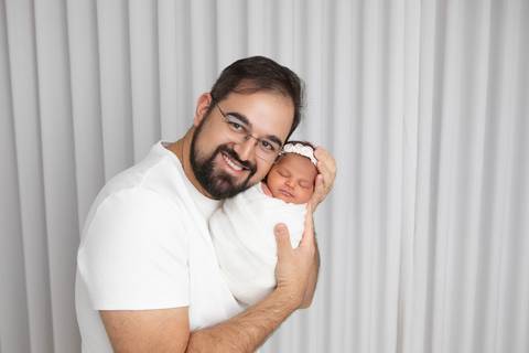 Proteção e cuidado paterno com a filha. O pai segurando a pequena Ísis nos braços em um momento de puro carinho e orgulho.'