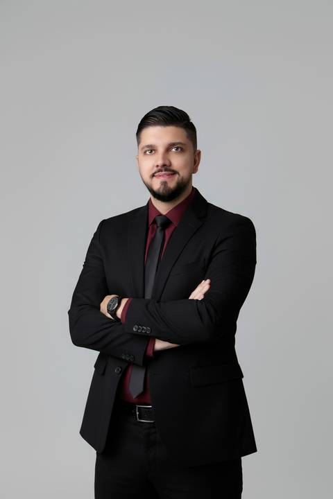Retrato de negócios masculino. Vitor com postura de segurança e braços cruzados em ensaio com terno escuro e camisa vinho.'