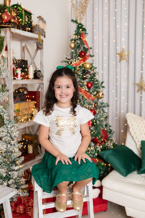 Pureza e expressões espontâneas na infância. Alana esbanjando alegria com blusa de laço dourado e saia verde em ambiente festivo protegido.'