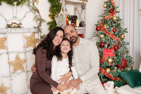Conexão e bem-estar em comemoração familiar. O casal e a filha Alana celebrando juntos com sorrisos espontâneos diante da árvore de Natal.'