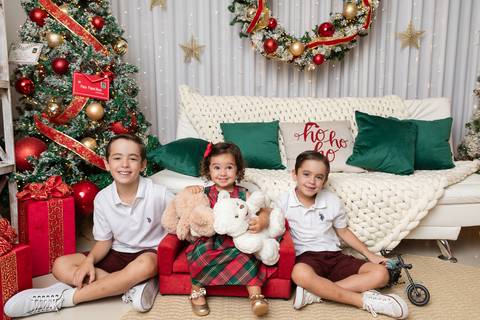 Interação espontânea e cumplicidade entre irmãos no cenário natalino. Davi, Lucas e Beatriz se divertindo juntos em ambiente infantil acolhedor em Brasília.'