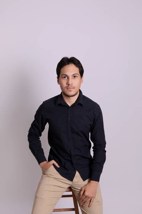 Retrato profissional masculino em estúdio. Christopher Dantas em pose casual com camisa escura e calça caqui em cenário minimalista claro.'
