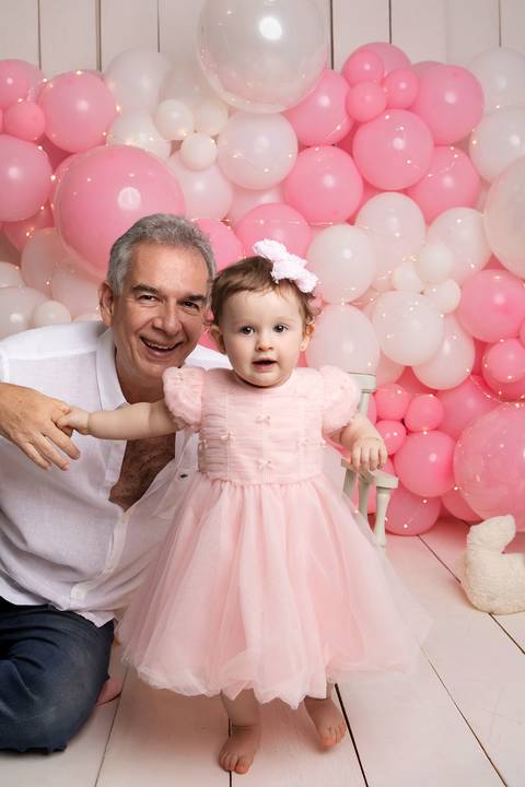 Cumplicidade e cuidado do pai com a bebê de um ano. Marcelo posando com a filha Maria Helena em cenário de festa.'