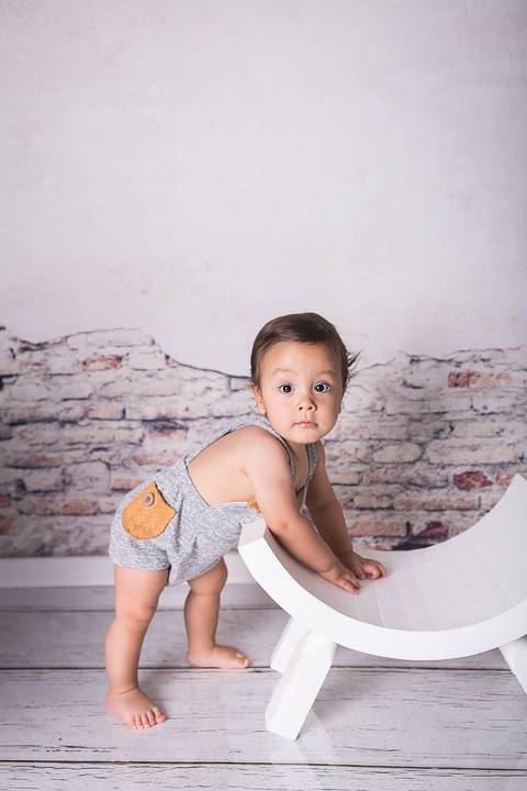 acompanhamento bebe book preço valor investimento brasilia df noroeste 10 meses fundo muro descascado vitor vestido macacão cinza prop cadeira banco curvado sorrindo mão no joelho em pé segurando'