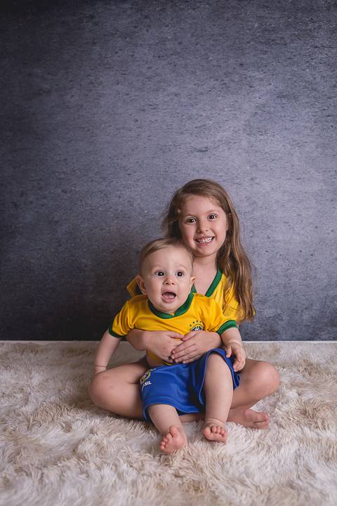 bebe book ensaio acompanhamento 06 07 meses fundo azul chão madeira caixinha de coração roupa azul de capuz sorrindo bebe sério tapete com a mamãe jeans e blusa branca irmã uniforme seleção brasileira futebol copa do mundo'
