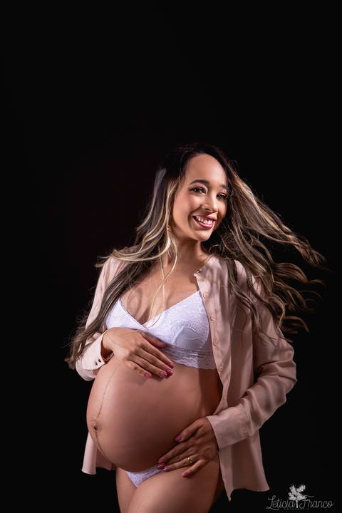 ensaio gestante mamãe fotografa fotógrafa brasília df distrito federal baby bebe reborn book melhor leticia franco letícia estúdio  gestante com a mão no cabelo e na barriga ligerie branca fundo preto sensual hobe casaco bege jogando o cabelo sorrindo'