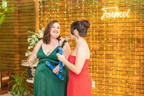 ALt text: a foto mostra uma formanda falando no microfone vestindo um vestido vermelho seus cabelos estão presos em um coque e são castanhos, enquanto a outra formanda sorri ela veste um vestido longo verde e também tem cabelos castanhos'