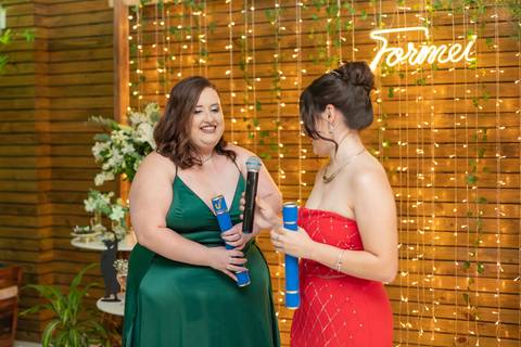 ALt text: a foto mostra uma formanda falando no microfone vestindo um vestido vermelho seus cabelos estão presos em um coque e são castanhos, enquanto a outra formanda sorri ela veste um vestido longo verde e também tem cabelos castanhos'